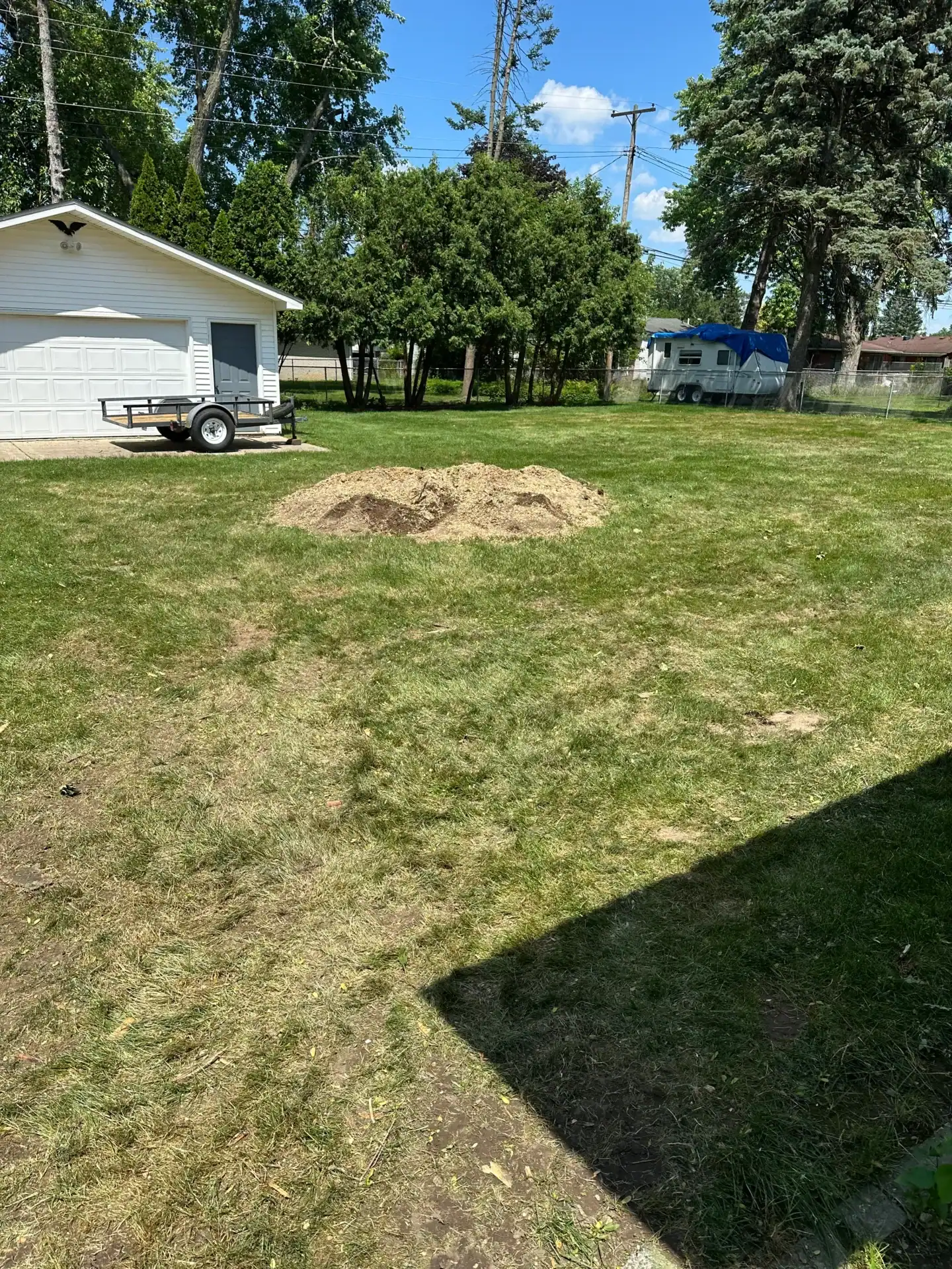 A pile of dirt rests on a sunny lawn near a garage, trailer, trees, and a Tree Services Oakland sign.