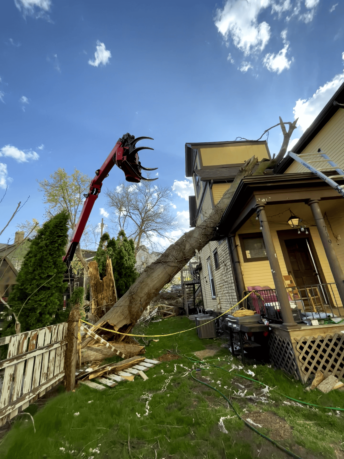 A crane removes a large fallen tree from an Oakland house, causing visible yard and fence damage.