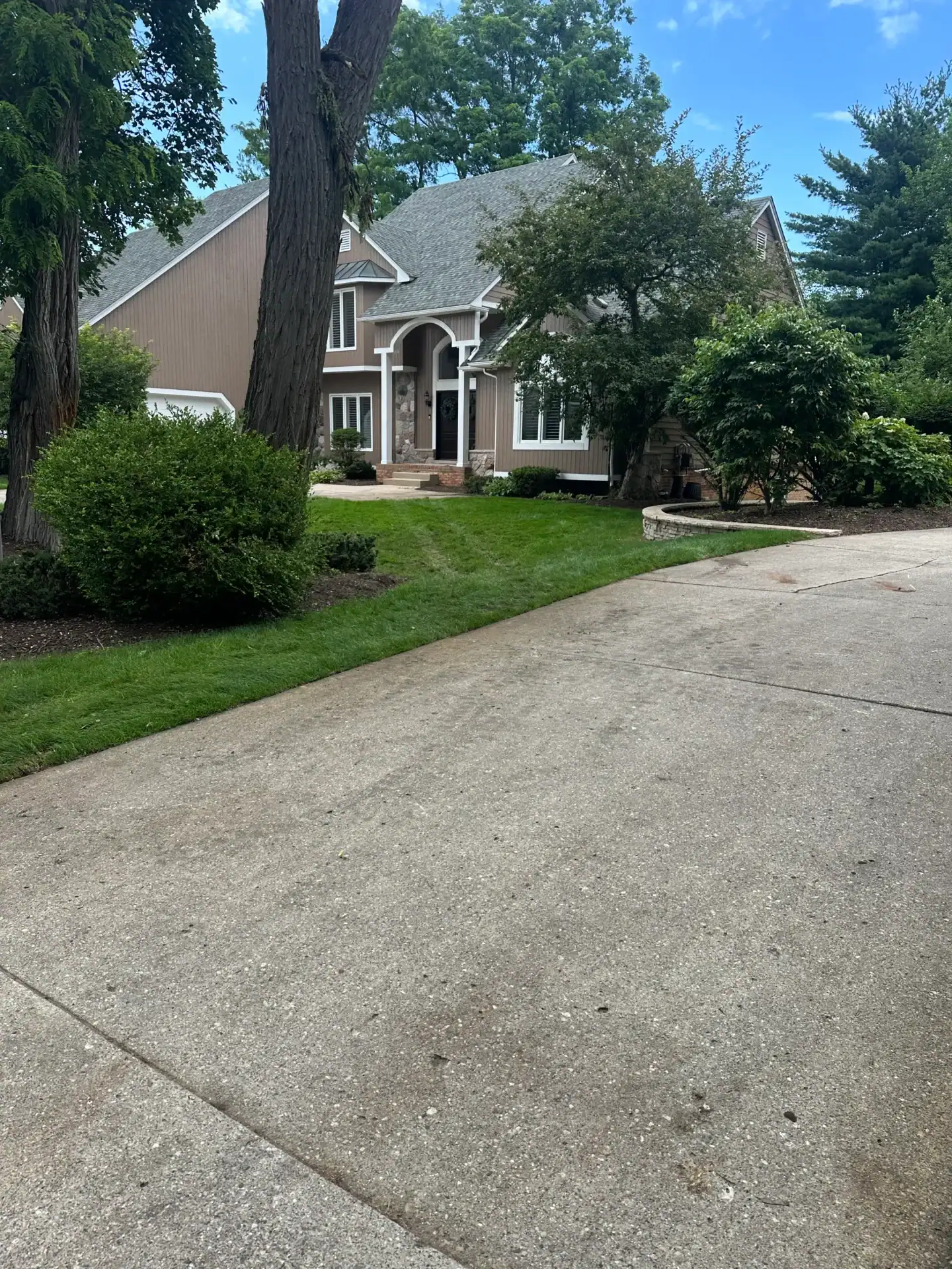 Concrete driveway to a brown MI house with white trim, surrounded by trees and green bushes.