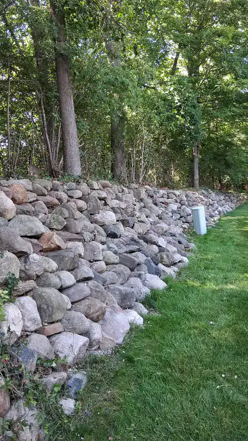A stone retaining wall slopes beside a grassy area with trees and a utility box in the background.