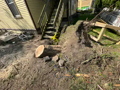 A tree stump and fallen tree rest by a tilted staircase in a dirt-filled yard next to a house.