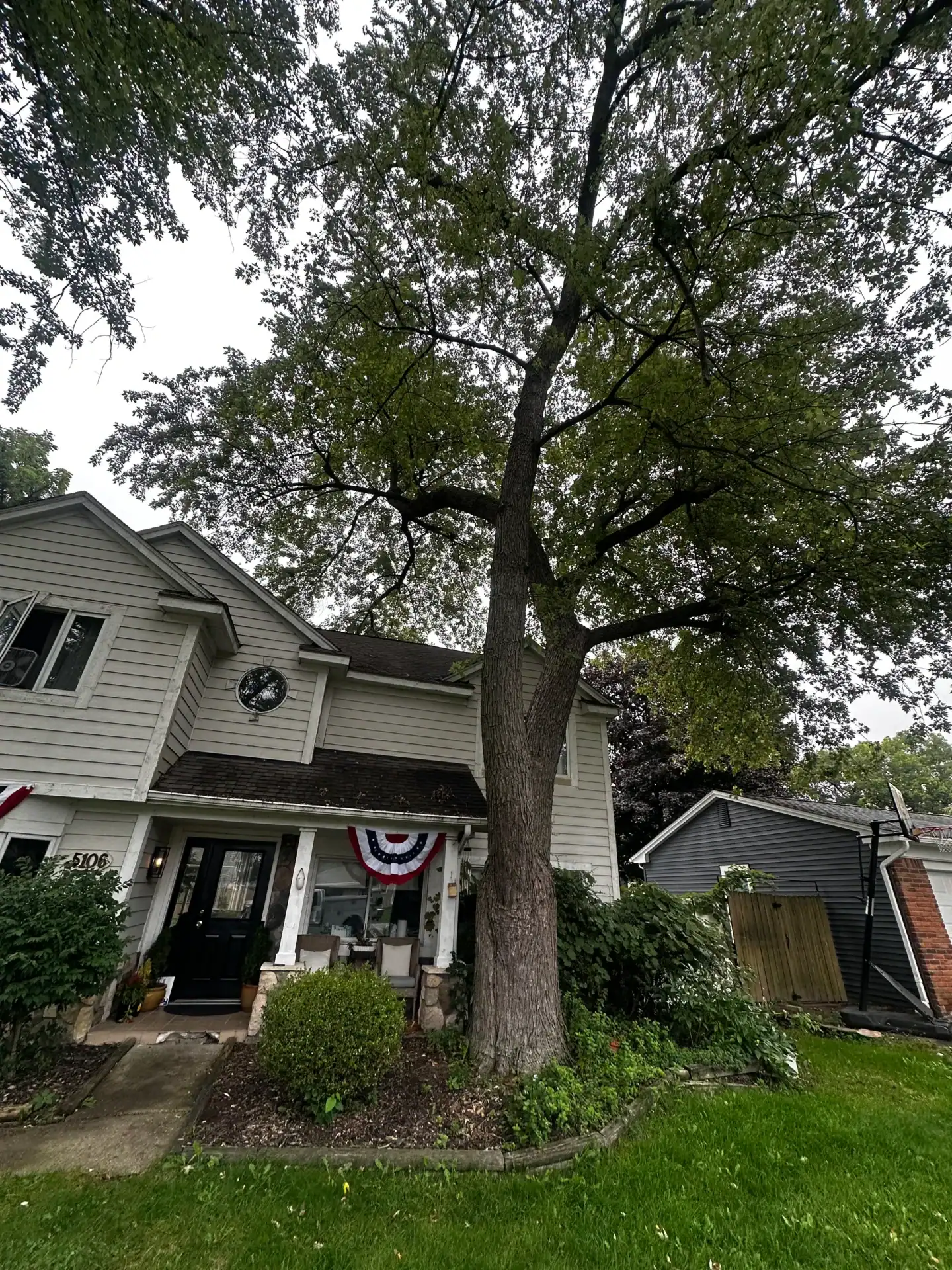 Two-story house with a green lawn, large front tree, and patriotic bunting on a cloudy afternoon.